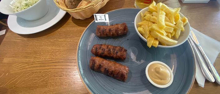 A plate of three grilled Romanian mici, skinless minced-meat rolls with a dark charred surface, served with chips, a pot of mustard, a bowl of coleslaw, a bread basket and a glass of beer on a wooden table.