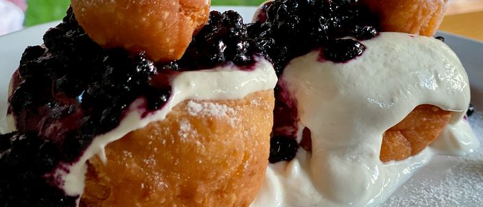 A close-up of two Romanian papanasi, fried cheese doughnuts, each a golden ring topped with a small dough ball, dusted with icing sugar and covered with white sour cream and dark berry jam.