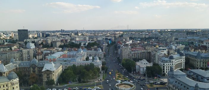Aerial daytime view over central Bucharest, a broad boulevard receding to a hazy horizon between rooftops of old and modern buildings, with a traffic roundabout and green trees in the foreground