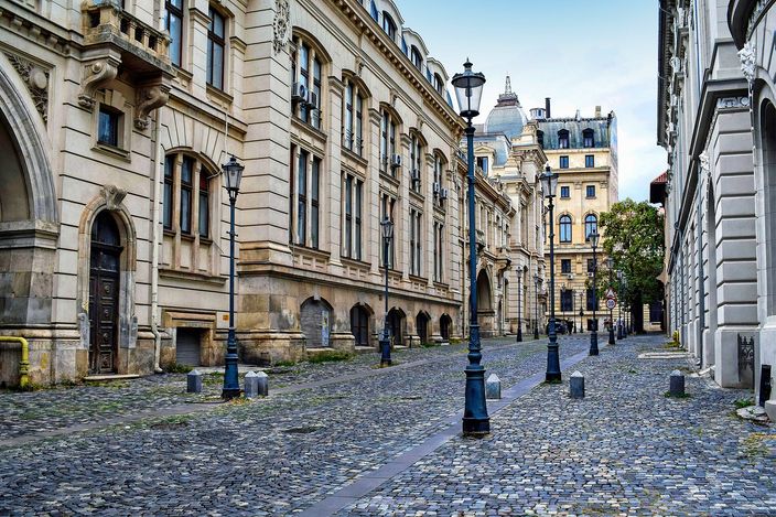 Empty cobbled street in Bucharest's Old Town, flanked by ornate stone 19th-century buildings with arched doorways and rows of cast-iron lampposts, a domed building closing the view