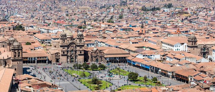 Cusco's Plaza de Armas seen from above in bright sun: the cathedral and the twin-towered church of La Compañía facing a green square, ringed by arcaded colonnades, with terracotta rooftops packed around it and the city climbing the steep hillside behind