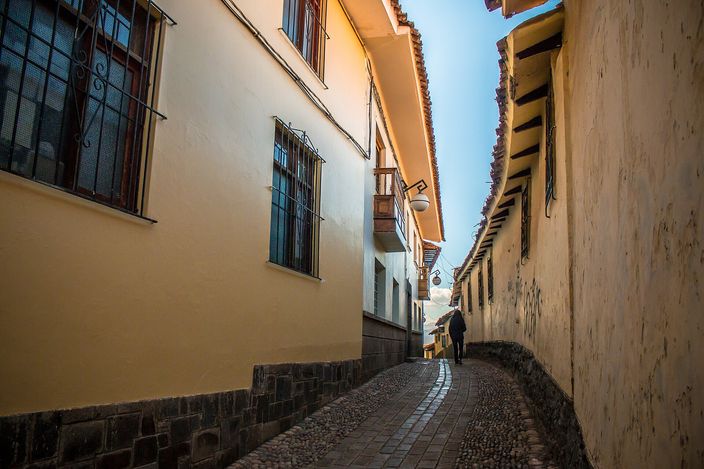 A narrow cobbled lane in Cusco's San Blas quarter climbing between whitewashed and ochre walls under tiled eaves, a lone figure walking up it in the shade