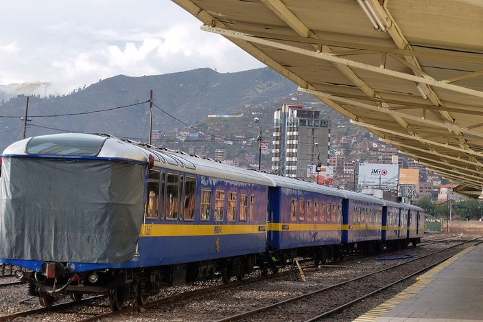 A blue and gold PeruRail train standing at a covered platform in Cusco, its lead carriage under a grey tarpaulin, with the city's houses covering the hillside behind