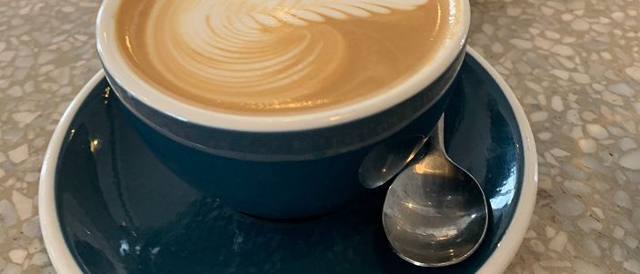 A flat white in a dark blue ceramic cup and saucer with a spoon, its surface a flat, glossy layer of microfoam holding a cleanly poured rosetta of latte art.