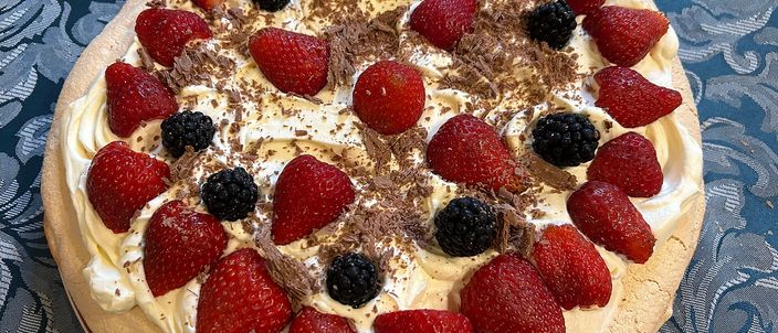 A whole round pavlova topped with softly whipped cream, fresh strawberries and blackberries and a scatter of chocolate shavings, set on a blue tablecloth.