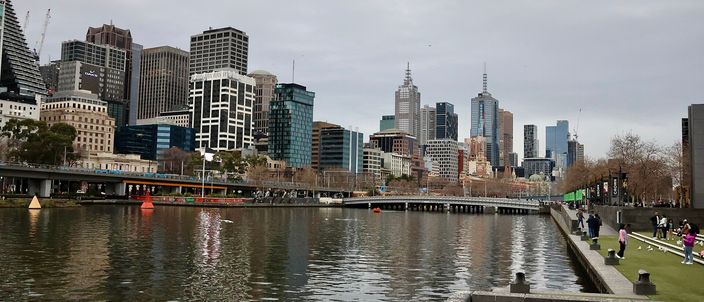 The Yarra River in the foreground with the Melbourne CBD skyline rising behind it under grey cloud, riverside promenade with walkers on the right, Flinders Street area visible
