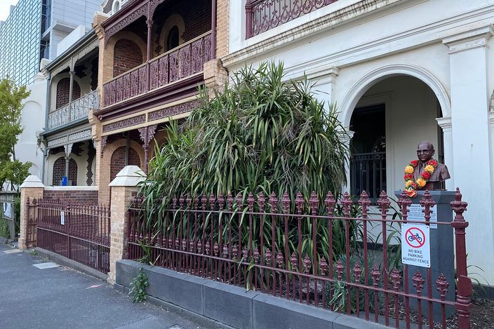 Ornate two-storey Victorian terrace houses with cast-iron lacework balconies and a low iron picket fence along a Carlton footpath, a modern tower rising behind