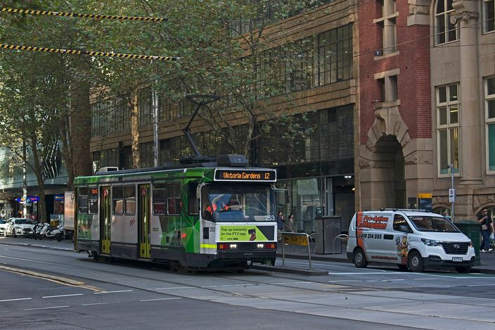 A green and yellow Melbourne tram signed 'Victoria Gardens' route 12 on Collins Street, overhead wires above, a white van and heritage buildings behind