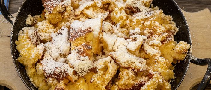 Torn pieces of Kaiserschmarrn in a black iron pan under a heavy dusting of icing sugar