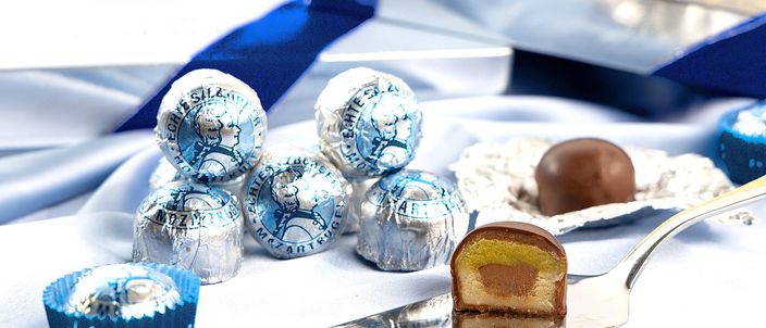 Several Mozartkugel wrapped in silver-and-blue foil on blue satin, with one unwrapped and cut in half on a spoon, showing the pale marzipan core, nougat layer and dark-chocolate coating.