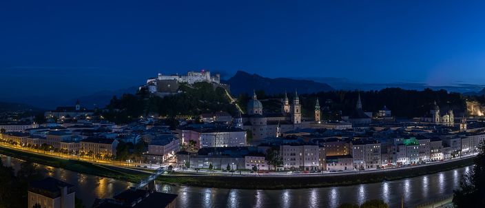Salzburg's old town at blue-hour dusk seen from the Kapuzinerberg: Hohensalzburg fortress floodlit on its rock above, the cathedral and church domes and towers, and the Salzach river reflecting lights in the foreground.