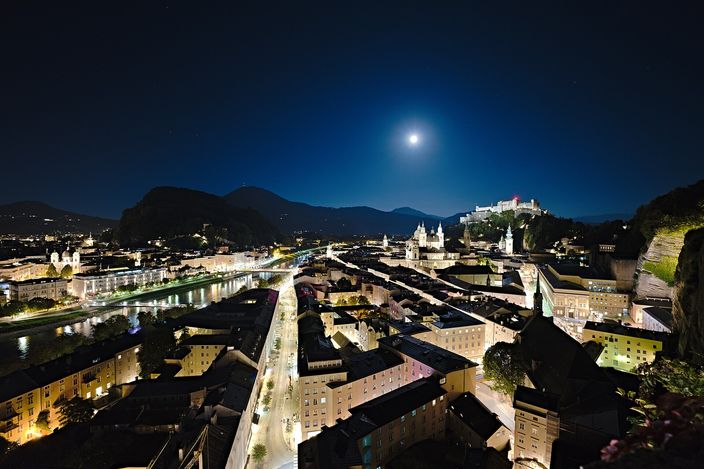 Night view over Salzburg's historic centre from the Mönchsberg: the fortress and cathedral domes lit up, the Salzach river curving through baroque rooftops, moon over the surrounding hills