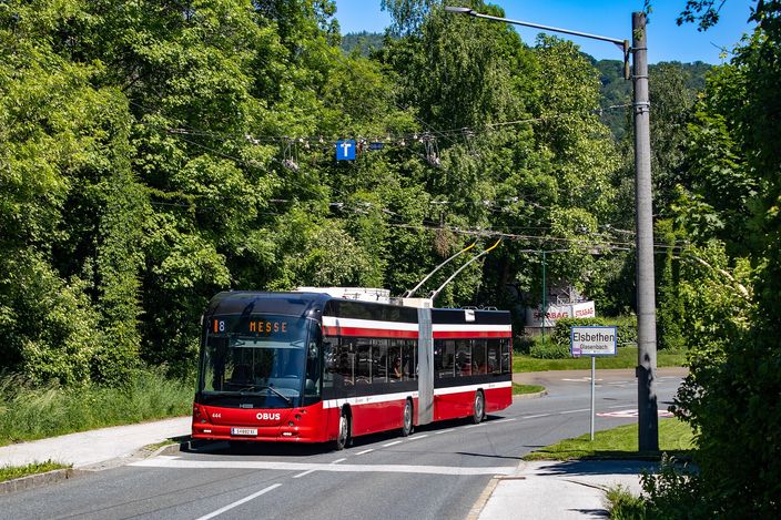 A red-and-black articulated Obus trolleybus on route 8 to Messe, drawing power from overhead wires on a tree-lined road at the city's edge, an Elsbethen place-name sign to the right