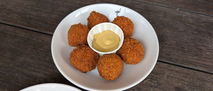 A white plate of golden-brown deep-fried bitterballen arranged around a small pot of yellow Dijon mustard, set on a dark wooden table.