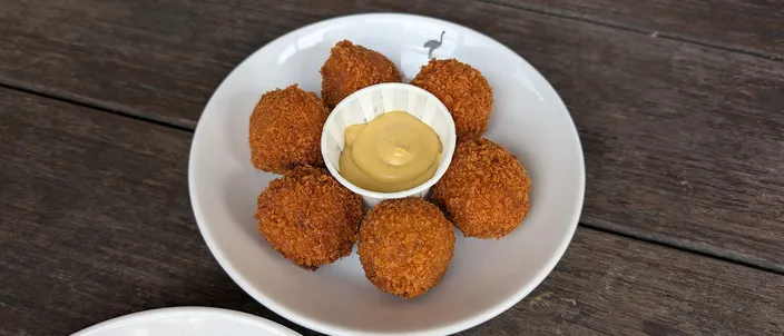 A white plate of golden-brown deep-fried bitterballen arranged around a small pot of yellow Dijon mustard, set on a dark wooden table.