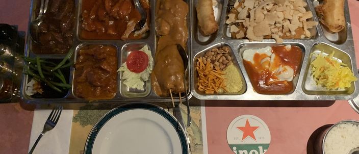 A rijsttafel spread laid out in metal serving trays and small bowls — rice, skewered satay in peanut sauce, and several curry and vegetable dishes arranged together across a table.