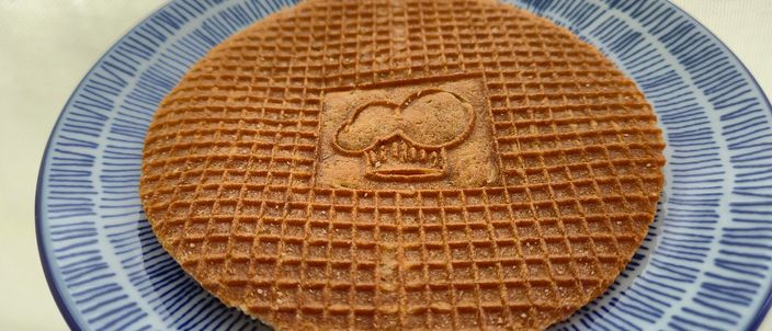 A single round stroopwafel — a lattice-patterned caramel-syrup waffle with a motif embossed into the top wafer — resting on a blue-and-white Dutch plate.