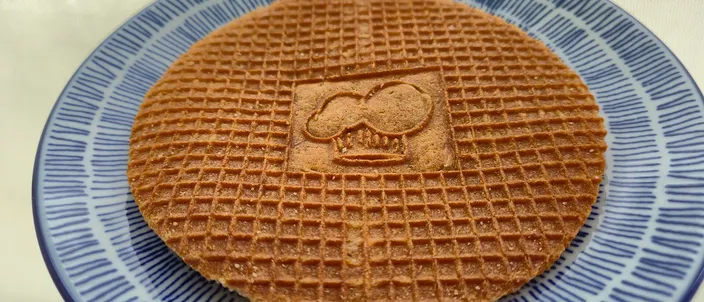 A single round stroopwafel — a lattice-patterned caramel-syrup waffle with a motif embossed into the top wafer — resting on a blue-and-white Dutch plate.