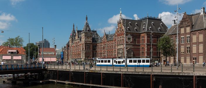 Amsterdam Centraal Station's red-brick neo-Gothic facade seen across the open water of the Open Havenfront, with two white-and-blue GVB trams drawn up on the quay in front of it and a small varnished canal boat passing in the foreground under a blue summer sky.