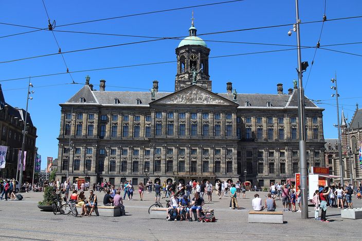 The long classical stone facade of the Royal Palace of Amsterdam on Dam Square under a clear blue sky, its central dome and cupola above the pediment, tram overhead wires crossing the frame, and people sitting and walking across the open square in front of it.
