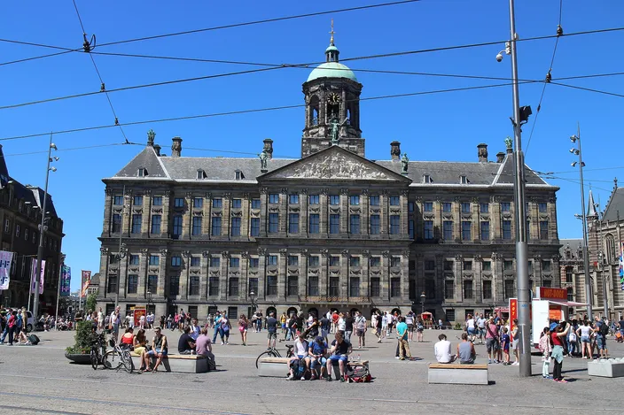The long classical stone facade of the Royal Palace of Amsterdam on Dam Square under a clear blue sky, its central dome and cupola above the pediment, tram overhead wires crossing the frame, and people sitting and walking across the open square in front of it.
