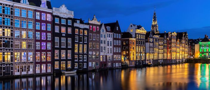A row of narrow, leaning gabled canal houses along the Damrak at blue hour, their windows lit yellow, pink and blue, mirrored in the still dark water in front of them, with a floodlit church tower rising behind the rooftops.