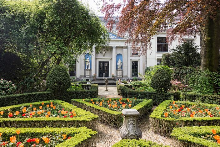 A formal town garden with low clipped box hedges enclosing beds of orange and yellow tulips, a stone urn on the central path, and a white neoclassical facade with statues in niches closing the far end under bare and blossoming trees.