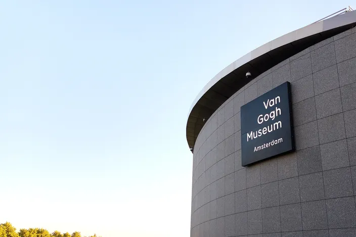 The curved gray stone drum of the Van Gogh Museum's elliptical wing against a clear pale sky at low sun, the museum's dark name plaque mounted on it and treetops along the bottom edge of the frame.