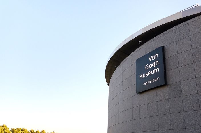 The curved grey stone drum of the Van Gogh Museum's elliptical wing against a clear pale sky at low sun, the museum's dark name plaque mounted on it and treetops along the bottom edge of the frame.