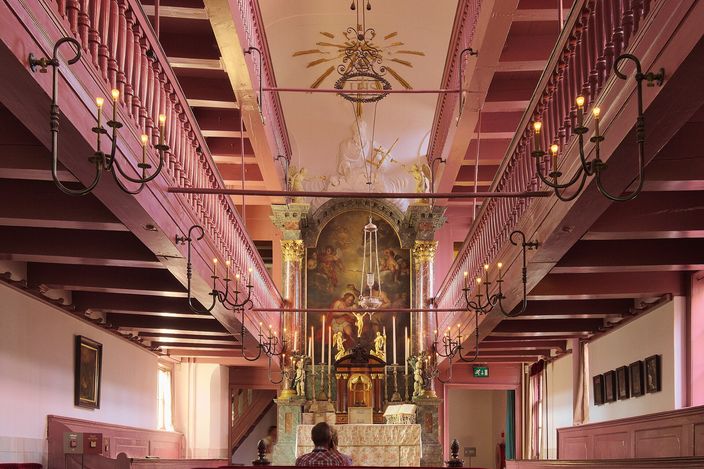 The interior of a hidden attic church: two tiers of pink-painted wooden galleries running down both side walls under a beamed ceiling, wrought-iron candle sconces along them, and a marble altar with a large painting above it at the far end, one seated visitor below.