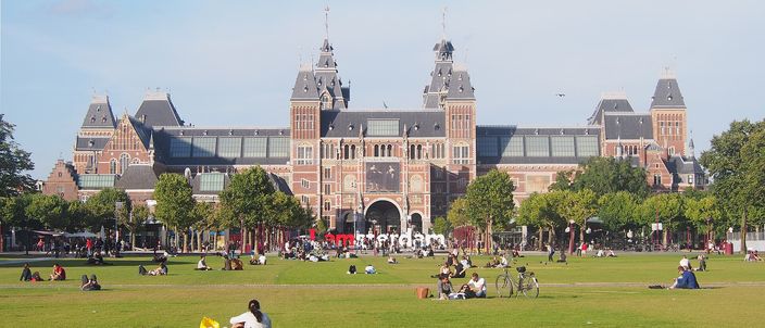 The long brick-and-stone facade of the Rijksmuseum with its twin towers and gabled centre seen across the wide open lawn of Museumplein on a clear summer day, people sitting on the grass and cycling along the paths in front of it.