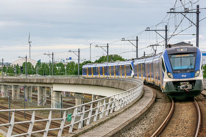A blue-and-white NS Sprinter train curving towards the camera on elevated track at Amsterdam Sloterdijk, overhead wires above it, a second train crossing on the viaduct behind and wind turbines on the flat skyline.