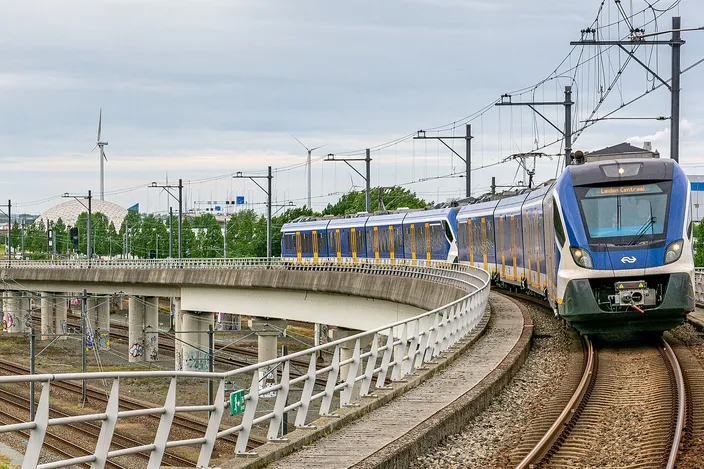 A blue-and-white NS Sprinter train curving towards the camera on elevated track at Amsterdam Sloterdijk, overhead wires above it, a second train crossing on the viaduct behind and wind turbines on the flat skyline.