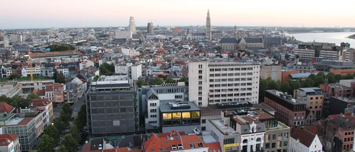 Rooftop view over Antwerp's old core at dusk: dense red-tiled roofs, the tall Gothic cathedral spire rising center-right, the river visible at the right edge under a pink sky