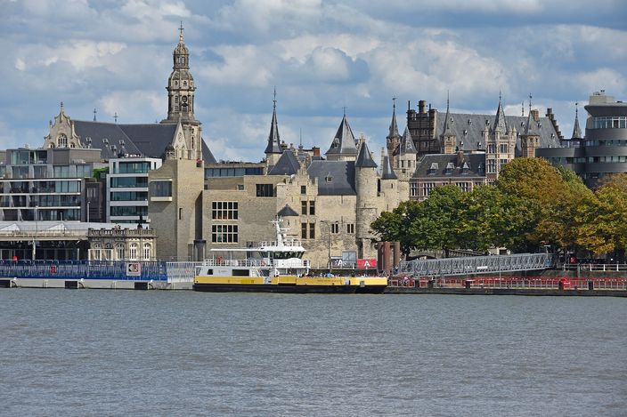 The crenellated grey stone gatehouse of Het Steen on the Antwerp quayside seen across the wide Scheldt from the opposite bank, with a tall clocktower and steep gabled roofs rising behind it, a yellow-and-white passenger boat moored below and autumn trees to the right.