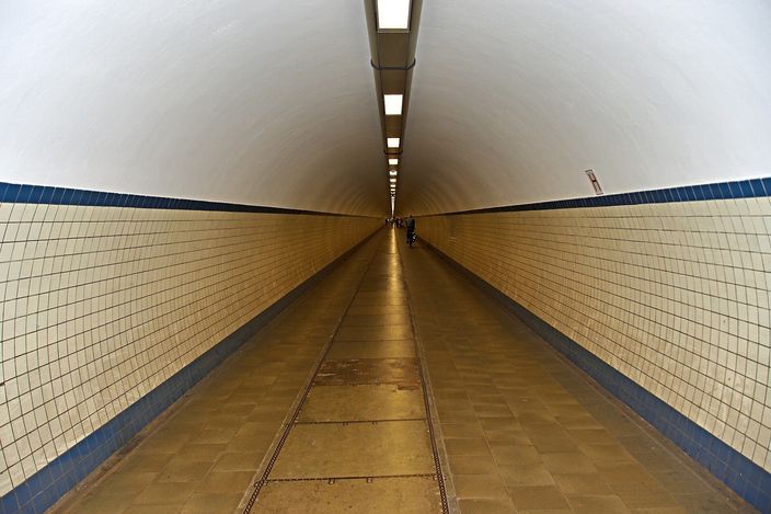 The interior of the Sint-Annatunnel under the Scheldt in Antwerp: a long white-and-cream tiled tube receding to a distant vanishing point, a blue tile band running along both walls at waist height, a strip of ceiling lights overhead and one small figure far down the passage.