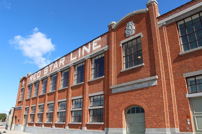 A long red-brick dockside shed in Antwerp with tall multi-paned steel windows and RED STAR LINE painted in large white capitals along its upper wall, a white anchor-and-star crest set into the curved end gable, under a clear blue sky.