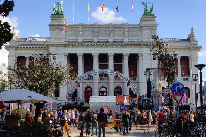 The white neoclassical facade of the Royal Museum of Fine Arts (KMSKA) in Antwerp's Zuid district, with bronze winged-horse sculptures on the roofline, flags, and a lively crowd, market stalls and parked bicycles filling the square below.