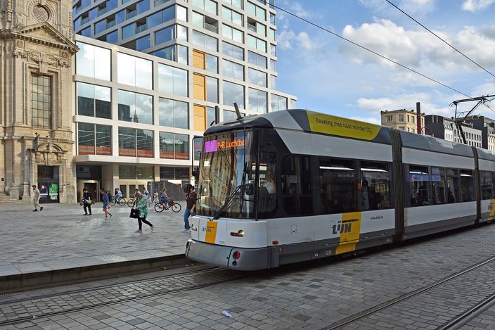 A white-and-yellow De Lijn tram signed route 1 to P+R Luchtbal running along street tracks past a baroque church facade and a modern glass building in central Antwerp, with cyclists and pedestrians crossing behind it in daylight.
