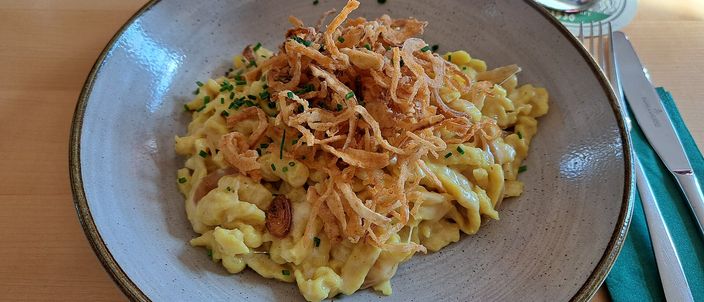 A bowl of golden Käsespätzle topped with dark crisp fried onions and chives, a beer glass and cutlery at the edge of the wooden table.