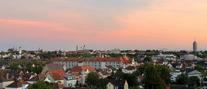 Augsburg's old town at dusk: red-tiled rooftops and church towers under a pink sky, a tall cylindrical tower rising at the right edge.