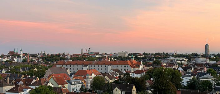 Augsburg's old town at dusk: red-tiled rooftops and church towers under a pink sky, a tall cylindrical tower rising at the right edge.