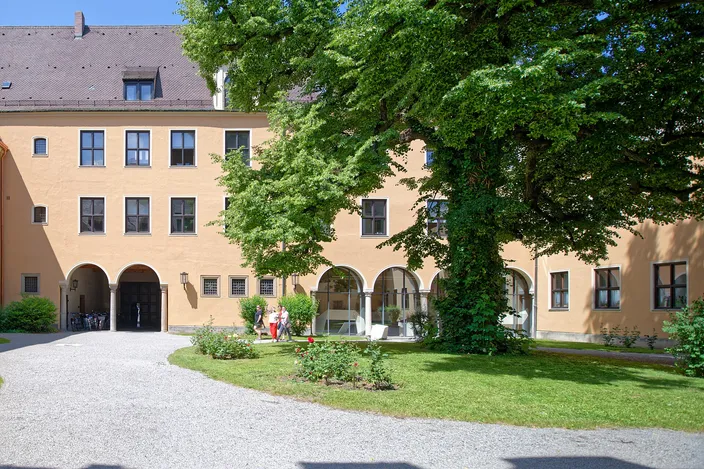 A sunlit courtyard of the Fugger houses in Augsburg: an ochre range with a ground-floor arcade of round arches, a large tree in leaf and a lawn with a rose bed in front of it.