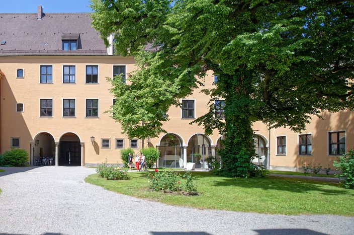 A sunlit courtyard of the Fugger houses in Augsburg: an ochre range with a ground-floor arcade of round arches, a large tree in leaf and a lawn with a rose bed in front of it.