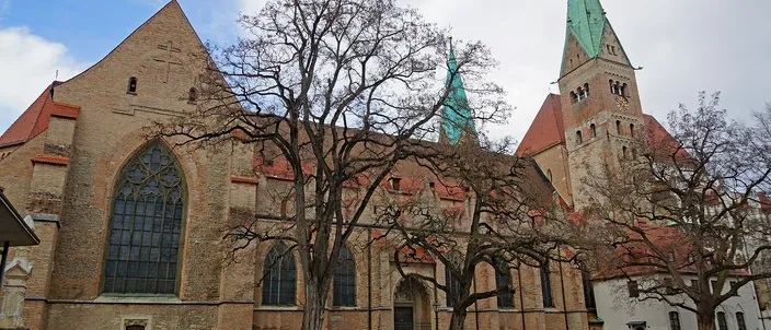 The long brick flank of Augsburg Cathedral seen across a cobbled square, bare winter trees in front of it and a tower with a green copper spire rising behind at the right.