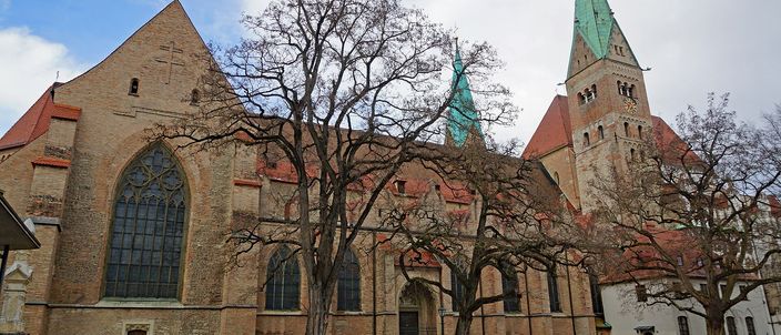 The long brick flank of Augsburg Cathedral seen across a cobbled square, bare winter trees in front of it and a tower with a green copper spire rising behind at the right.
