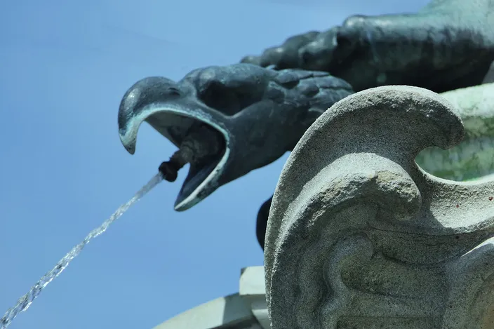 A bronze eagle-headed spout on the Mercury fountain in Augsburg, mouth open, a jet of water arcing out of it against a clear blue sky, weathered green metal beside pale carved stone