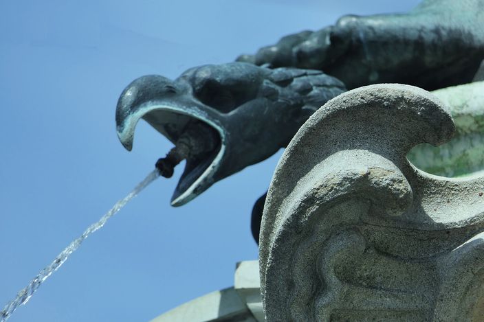 A bronze eagle-headed spout on the Mercury fountain in Augsburg, mouth open, a jet of water arcing out of it against a clear blue sky, weathered green metal beside pale carved stone