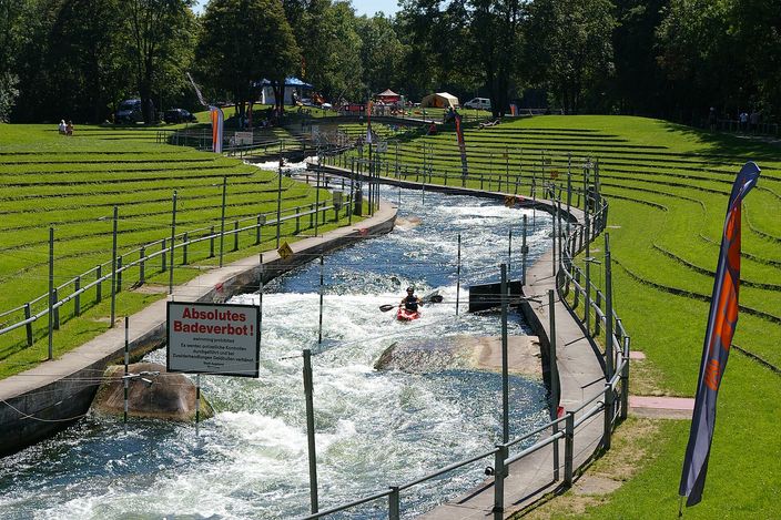 The Augsburg whitewater canal seen from above: a kayaker in the rapids between hanging slalom gates, terraced green banks on both sides, and a red and white sign in the foreground reading Absolutes Badeverbot