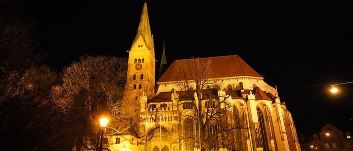 Augsburg cathedral at night, floodlit warm orange against a black sky, its Romanesque tower on the left and the Gothic east end on the right, bare trees and a streetlamp in front